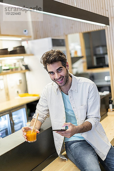 Porträt eines lachenden Mannes mit Handy  der an der Theke einer Bar sitzt und einen Drink nimmt