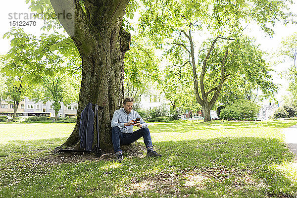Geschäftsmann mit E-Scooter lehnt mit Smartphone an Baumstamm im Stadtpark