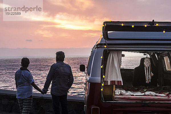 Älteres Ehepaar reist in einem Oldtimer-Van und betrachtet den Sonnenuntergang am Meer