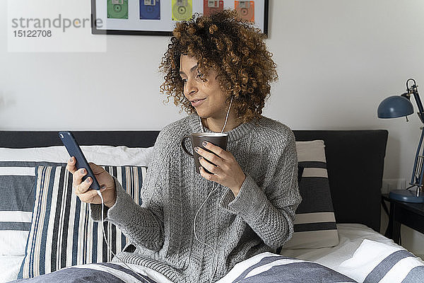 Frau sitzt im Bett  trinkt Kaffee  benutzt Smartphone und Kopfhörer