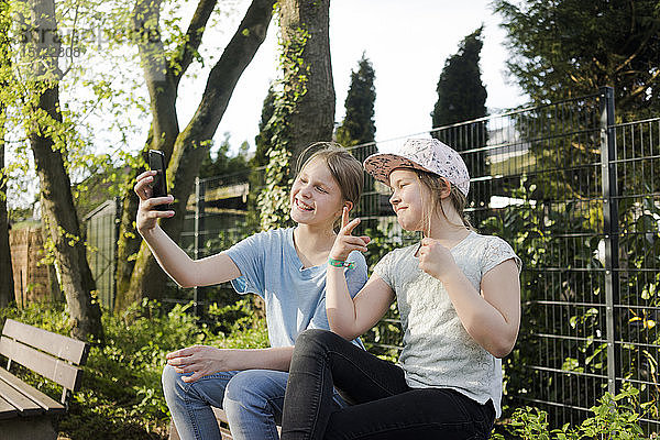 Zwei glückliche Mädchen sitzen auf einer Parkbank und machen ein Selfie