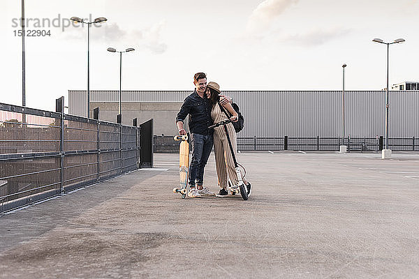 Junges Paar mit Longboard und Elektroroller umarmt sich auf dem Parkdeck