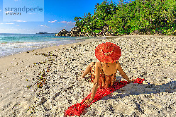 Touristin mit rotem Sonnenhut sitzt am unberührten weißen Strand bei Sonnenuntergang  Anse Georgette Beach  Praslin Island  Seychellen  Indischer Ozean  Afrika