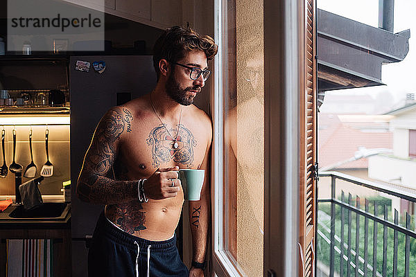 Mittelgroßer erwachsener Mann mit Tätowierungen  der durch ein Küchenfenster hinaussieht