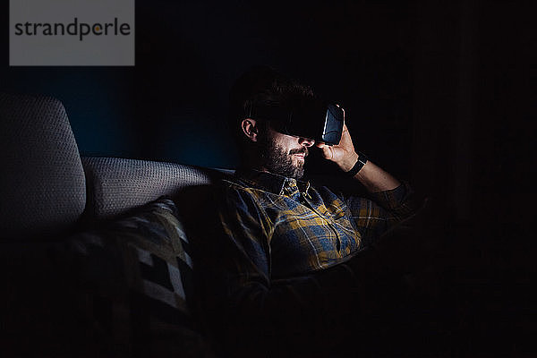 Bärtiger mittelgroßer Mann  der im Dunkeln auf dem Sofa sitzt und durch ein Virtual-Reality-Headset nach unten blickt