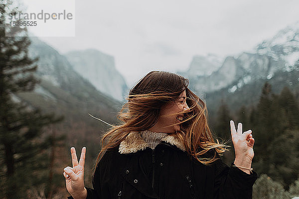 Junge Frau  die lange braune Haare schüttelt und ein Friedenszeichen in einer Berglandschaft setzt  Yosemite Village  Kalifornien  USA