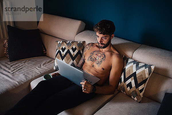 Mittelgroßer erwachsener Mann mit tätowierter Brust auf dem Sofa beim Blick auf den Laptop