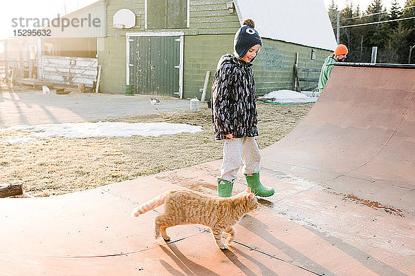 Junge mit Katze schaut auf die Skateboard-Rampe auf dem Bauernhof