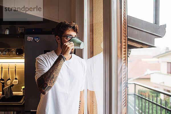 Mittelgroßer erwachsener Mann trinkt Kaffee  während er durch ein Wohnungsfenster hinausschaut