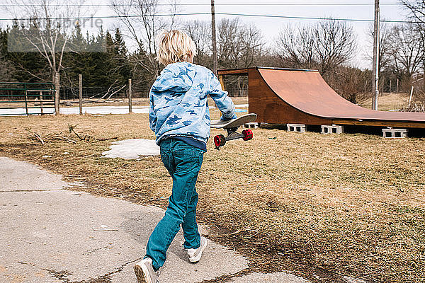 Blonder Junge trägt Skateboard zur ländlichen Skateboard-Rampe  Rückansicht