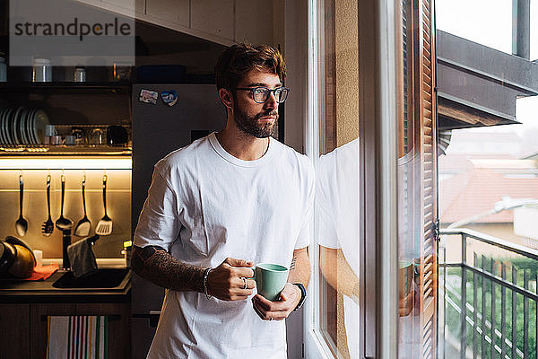 Mittelgroßer erwachsener Mann mit Kaffeetasse  der durch das Wohnungsfenster hinausblickt