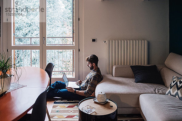 Mittelgroßer erwachsener Mann sitzt mit Laptop auf dem Wohnzimmerboden
