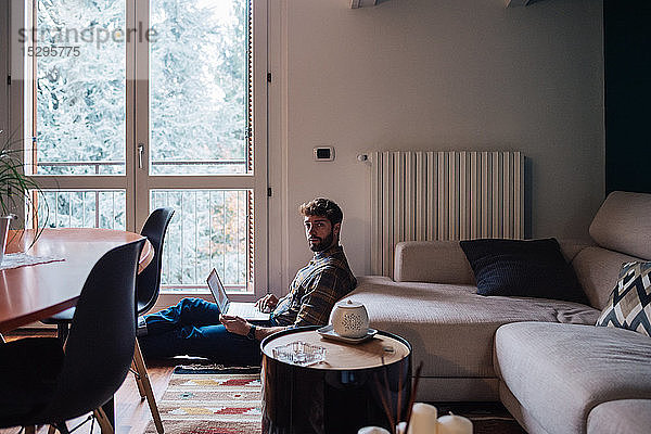 Mittelgroßer erwachsener Mann sitzt mit Laptop auf dem Wohnzimmerboden  Portrait