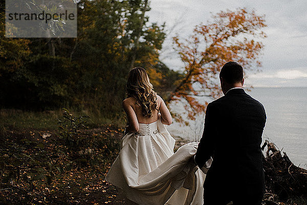 Romantischer junger Bräutigam trägt die Schleppe des Brautkleides am Seeufer  Rückansicht