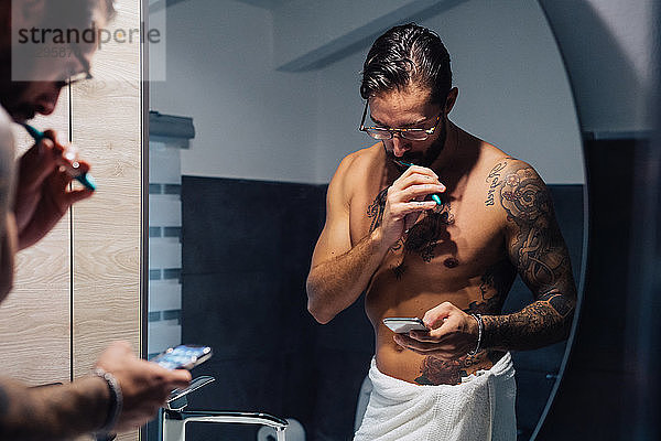 Mittelgroßer erwachsener Mann mit Tätowierungen  der sich die Zähne putzt und sein Smartphone am Badezimmerspiegel betrachtet  Spiegelbild