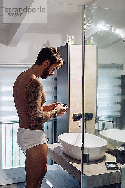 Mittelgroßer erwachsener Mann mit Tätowierungen  der sein Smartphone am Badezimmerspiegel betrachtet