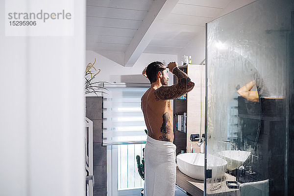 Mittelgroßer erwachsener Mann mit Tätowierungen  der am Badezimmerspiegel Haare kämmt