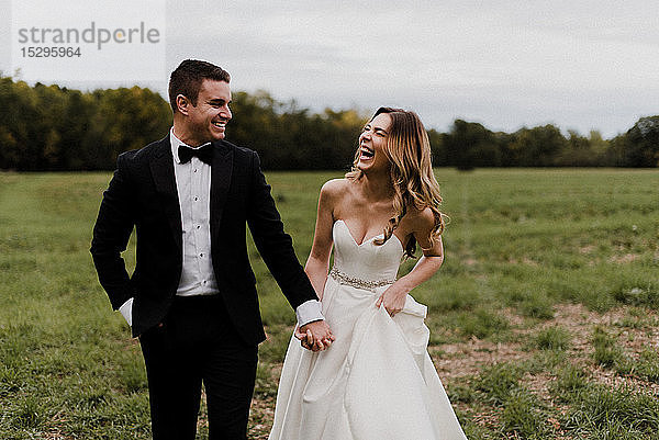 Romantische junge Braut und Bräutigam halten sich an den Händen und lachen auf dem Feld