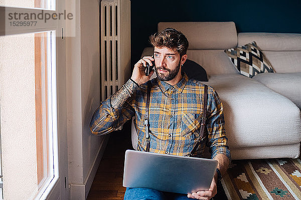 Mittelgroßer erwachsener Mann sitzt auf dem Wohnzimmerboden mit Laptop und telefoniert mit einem Smartphone