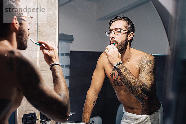 Mittelgroßer erwachsener Mann mit Tätowierungen beim Zähneputzen am Badezimmerspiegel