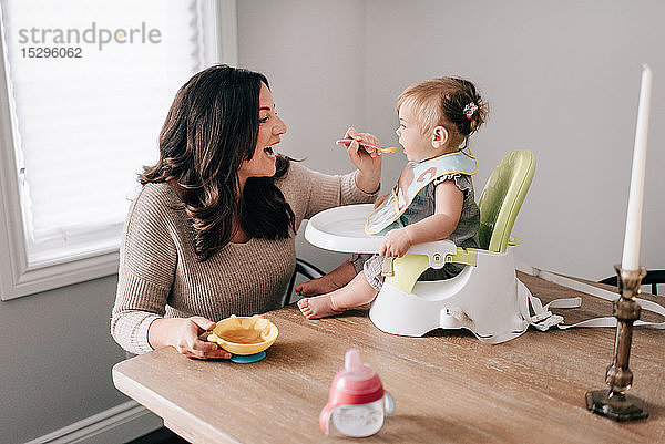 Mutter füttert Baby-Tochter im Kindersitz auf dem Küchentisch