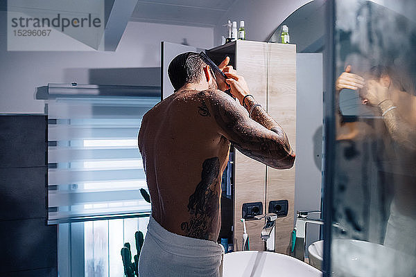 Mittelgroßer erwachsener Mann mit Tätowierungen  der am Badezimmerspiegel Haare kämmt