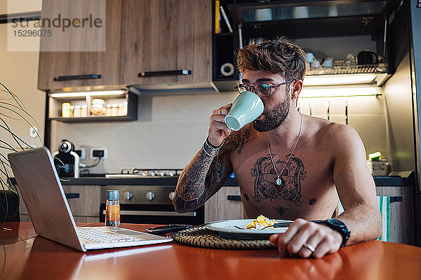 Mittelgroßer erwachsener Mann mit Tätowierungen  der Kaffee trinkt und dabei auf einen Laptop schaut