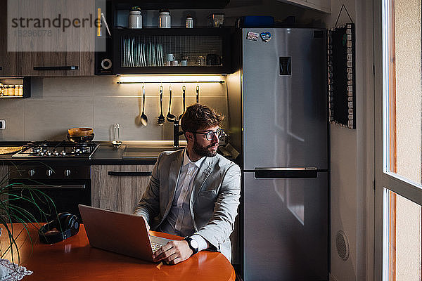 Mittelgroßer erwachsener Mann sitzt am Küchentisch mit Laptop und schaut aus dem Fenster