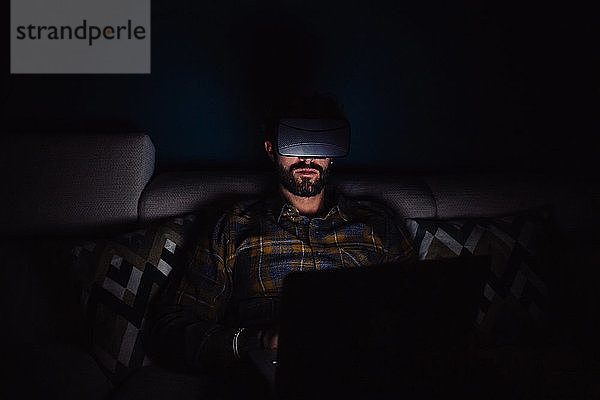 Bärtiger mittelgroßer Mann sitzt im Dunkeln auf dem Sofa und schaut durch ein Virtual-Reality-Headset
