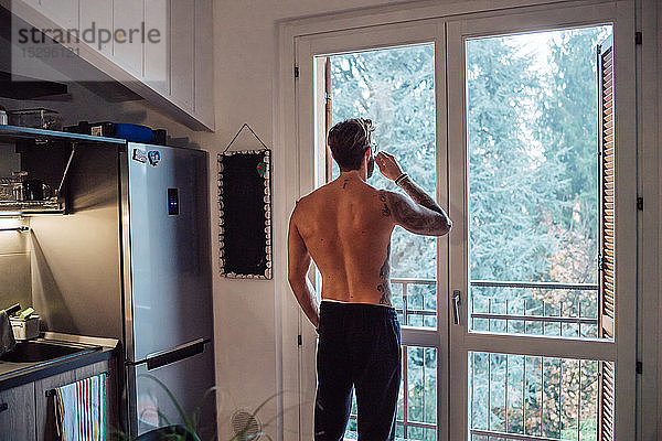 Mittelgroßer erwachsener Mann mit Tätowierungen  der Kaffee trinkt und durch ein Küchenfenster hinaussieht