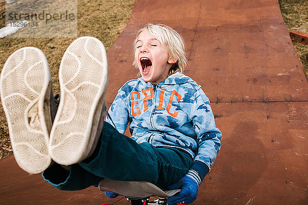 Junge sitzend und Skateboard fahrend rückwärts die hölzerne Skateboard-Rampe hinunter
