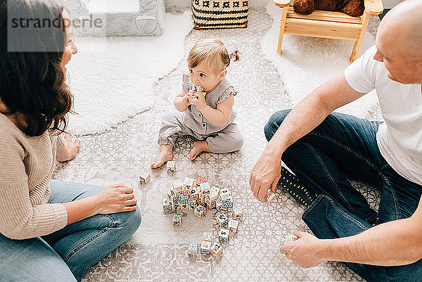 Mutter und Vater sitzen auf dem Kinderzimmerboden  während die kleine Tochter mit Bauklötzen spielt