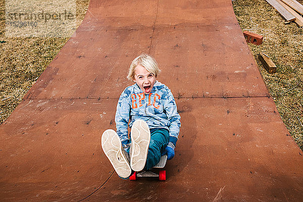 Junge sitzend und Skateboard fahrend rückwärts die hölzerne Skateboardrampe hinunter  Portrait
