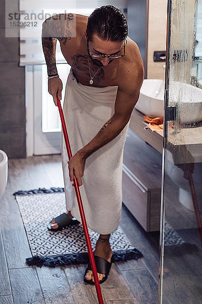 Mittelgroßer erwachsener Mann in Handtuch gewickelt reinigt Badezimmerboden