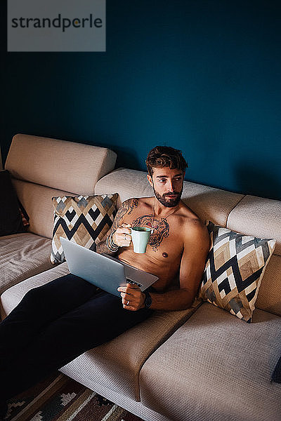 Mittelgroßer erwachsener Mann mit tätowierter Brust auf Sofa mit Laptop und Kaffee