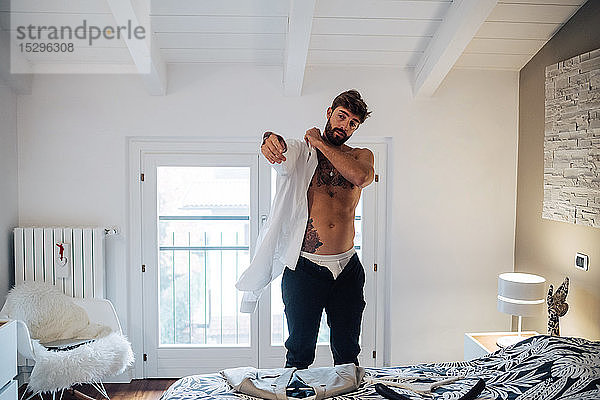 Mittelgroßer erwachsener Mann mit Tätowierungen  der sich im Schlafzimmer ein Hemd anzieht