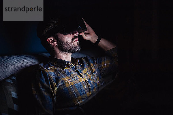 Bärtiger mittelgroßer Mann  der im Dunkeln auf dem Sofa sitzt und durch ein Virtual-Reality-Headset nach oben schaut