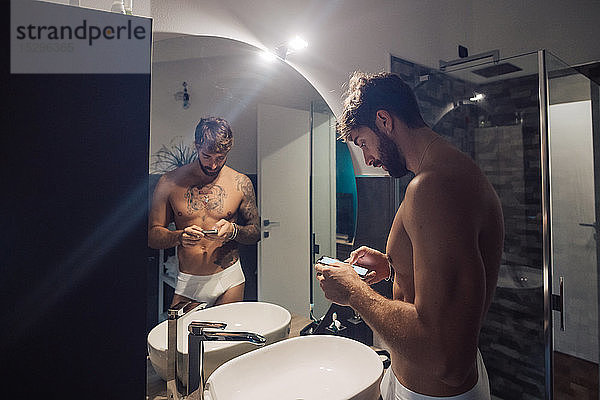 Mittelgroßer erwachsener Mann mit Tätowierungen über Smartphone-Touchscreen am Badezimmerspiegel  Spiegelbild