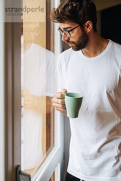 Mittelgroßer erwachsener Mann mit Kaffee  der durch ein Wohnungsfenster hinaussieht