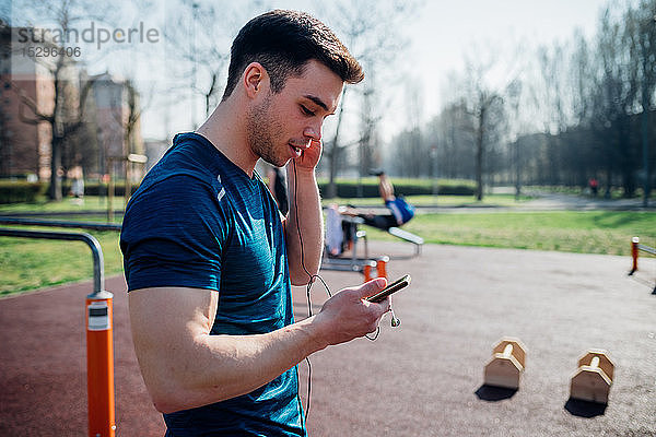 Gymnastik im Fitnessstudio im Freien  junger Mann hört Kopfhörer und schaut auf ein Smartphone