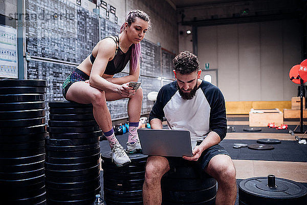 Junge Frau und Mann trainieren gemeinsam im Fitnessstudio  sitzen auf Gewichten und schauen auf den Laptop