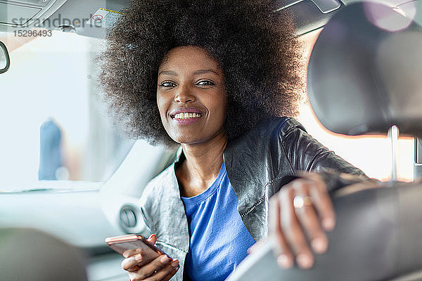 Glückliche junge Frau mit Afro-Haaren auf dem Beifahrersitz eines Autos  die ein Smartphone hält  Porträt