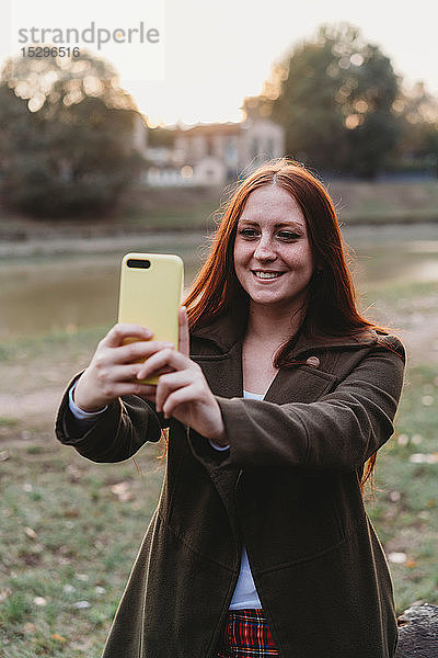 Junge Frau mit langen roten Haaren beim Smartphone-Selfie am Flussufer in der Abenddämmerung  Florenz  Toskana  Italien