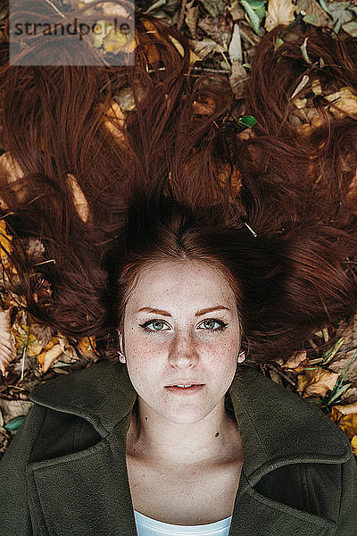 Junge Frau mit langen roten Haaren inmitten von Herbstblättern liegend  Überkopfporträt