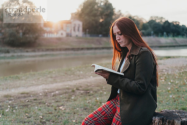 Junge Frau mit langen roten Haaren sitzt auf Baumstumpf und liest am Flussufer bei Sonnenuntergang ein Buch  Florenz  Toskana  Italien