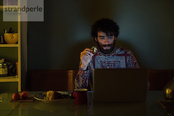 Bärtiger junger Mann frühstückt mit Laptop zu Hause