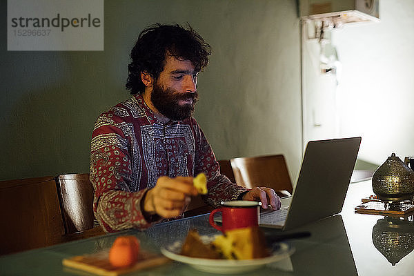 Bärtiger junger Mann frühstückt mit Laptop zu Hause