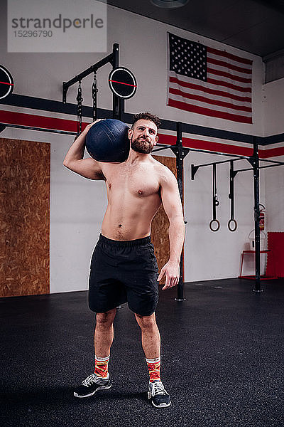 Junger Mann trainiert  trägt Atlasball auf der Schulter im Fitnessstudio
