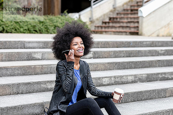 Glückliche junge Frau mit Afro-Haaren sitzt auf einer Stadttreppe und hört ein Smartphone