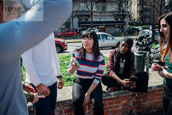 Junge weibliche und männliche erwachsene Freunde sitzen an der Wand  chatten und schauen auf Smartphones
